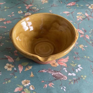 May include: A handmade, round, yellow-brown ceramic bowl with a pouring spout. The bowl has a smooth interior and a slightly textured exterior. The bowl is sitting on a teal floral patterned surface.