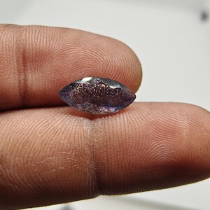 May include: A marquise-cut gemstone, deep purple in colour with sparkling inclusions. The gem is held between fingers, highlighting its shape and colour. The stone's facets reflect light, creating a brilliant display. This gemstone is suitable for jewellery making.