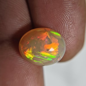 May include: An oval-shaped cabochon gemstone with a translucent, milky base color. The stone displays a vibrant play-of-color, featuring flashes of bright orange, neon green, and yellow light reflecting from within its smooth, polished surface.