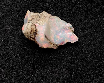 May include: A rough, uncut opal gemstone with a milky white base color and flashes of iridescent pink, blue, and orange. The opal is set against a dark, textured background, showcasing its natural beauty.