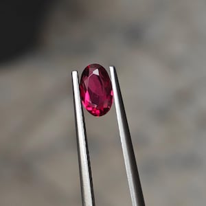 Könnte beinhalten: Nahaufnahme eines oval geschliffenen, leuchtend roten Rubins, gehalten von einer silbernen Pinzette. Die Facetten des Edelsteins fangen das Licht ein. Der Hintergrund ist ein weiches Grau, das die Brillanz des Rubins hervorhebt. Detailaufnahme des Edelsteins.