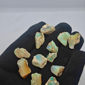 Puede incluir: Una colección de piedras preciosas de ópalo en bruto, que muestra una variedad de colores, incluyendo azul, verde y naranja. Los ópalos tienen un aspecto natural, sin pulir, con texturas y patrones visibles. Las piedras preciosas se muestran sobre un fondo oscuro.