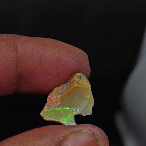 May include: A raw opal gemstone, displaying a spectrum of colours such as orange, green, and yellow. The stone is held between fingers against a dark backdrop, highlighting its natural, irregular form and iridescent colour play.
