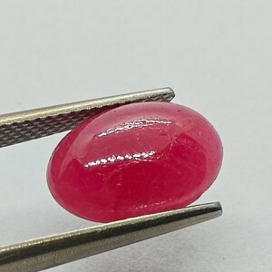 May include: A polished, oval-shaped, red gemstone held by a pair of silver tweezers. The gem has a smooth, glossy surface and a vibrant, saturated color. The tweezers are in sharp focus, highlighting the gem's shape and color.
