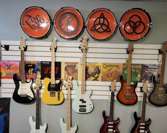 May include: A wall display of various electric guitars and basses, including a black Fender Stratocaster, a yellow Fender Telecaster, a light blue bass guitar, a brown sunburst guitar, and a black and brown guitar. The wall is decorated with vintage album covers and four round drumheads with logos, including "Zoso", a feather, a triquetra, and the "three circles" logo.