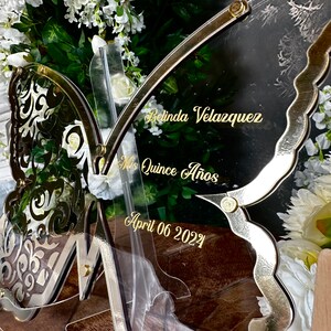 Puede incluir: Un letrero de acr&iacute;lico transparente en forma de mariposa con detalles dorados y el texto "Belinda Velazquez", "Mis Quince Anos" y "April 06 2022".