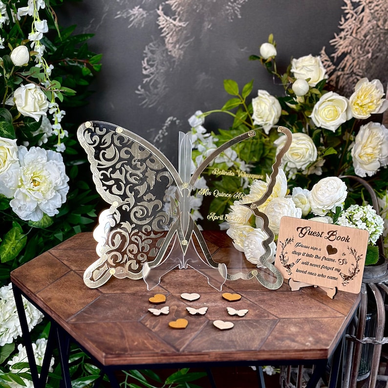 Puede incluir: Un libro de visitas de mariposa de acr&iacute;lico transparente con un dise&ntilde;o de filigrana dorada. La mariposa est&aacute; de pie sobre una base de madera. Un letrero de madera con el texto "Guest Book" est&aacute; en primer plano. El letrero tiene un dise&ntilde;o floral y el texto "Please sign a chip it into the frame. So I will never forget my loved ones who came."