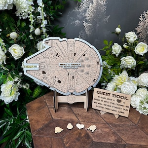 May include: A wooden guest book shaped like the Millennium Falcon from Star Wars. The ship is decorated with a silver finish and has a small wooden stand. The guest book has a sign that reads "Guest Book" and "Please sign a & drop it into the frame so we will never forget our loved ones who came."