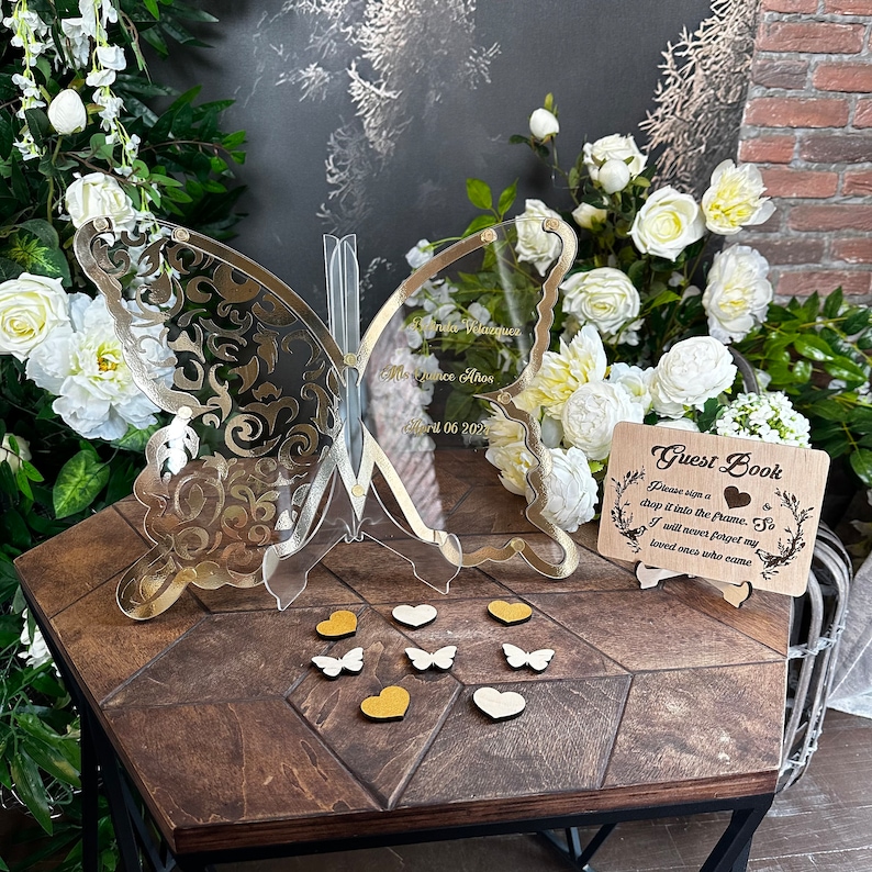 Puede incluir: Un libro de visitas de mariposa de acr&iacute;lico transparente con detalles dorados. La mariposa est&aacute; sobre una mesa de madera con un peque&ntilde;o letrero de madera que dice "Libro de visitas Por favor, firma un trozo y ponlo en el marco. As&iacute;, nunca olvidar&eacute; a mis seres queridos que vinieron".