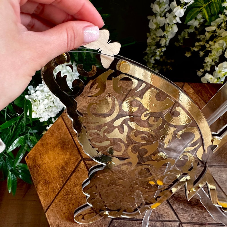 Puede incluir: Una mariposa de acr&iacute;lico transparente con un dise&ntilde;o dorado. La mariposa est&aacute; en un soporte y tiene una peque&ntilde;a mariposa de madera en la parte superior.