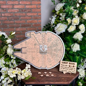 May include: A wooden guest book shaped like the Millennium Falcon from Star Wars. The guest book has a silver outline and a sign that says "GUEST BOOK Please sign a  or drop it into the frame. So we will never forget our loved ones who came."