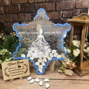 Pu&ograve; includere: Un libro degli ospiti a forma di fiocco di neve con un bordo blu e un design da sposa tagliato al laser. Viene visualizzato il testo "Jazlene Luz Guzman March 25th 2023". Include un cartello di legno "Guest Book" e piccoli cuori di legno.