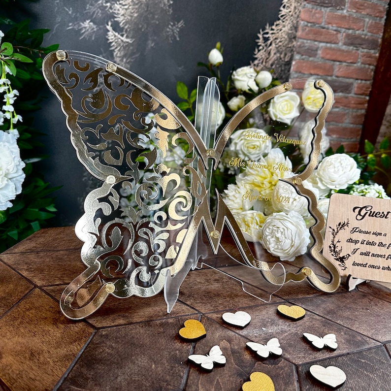 Puede incluir: Un libro de visitas en forma de mariposa de acr&iacute;lico transparente y dorado con un letrero que dice "Por favor, firme y d&eacute;jelo caer en el frasco. Nunca olvidar&eacute; a los seres queridos cuando mire este frasco."