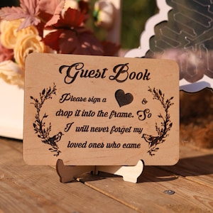 Pu&ograve; includere: Cartello per libro degli ospiti in legno con la scritta "Guest Book" in elegante corsivo. Il cartello include illustrazioni di cuori e uccelli, con il testo "Please sign a drop it into the frame. So I will never forget my loved ones who came."