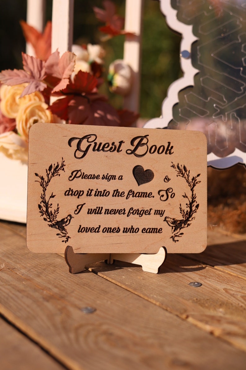 Pu&ograve; includere: Cartello per libro degli ospiti in legno con la scritta "Guest Book" in elegante corsivo. Il cartello include illustrazioni di cuori e uccelli, con il testo "Please sign a drop it into the frame. So I will never forget my loved ones who came."