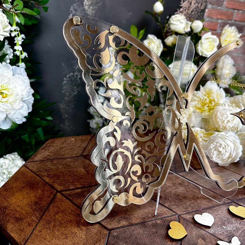 Puede incluir: Un letrero de acr&iacute;lico en forma de mariposa dorada con un patr&oacute;n de filigrana decorativo. El letrero dice "Belinda Velazquez" y "Mis Quince A&ntilde;os" con la fecha "Oct 06 2023".