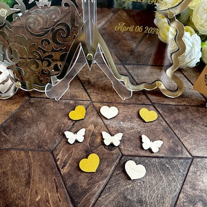 Puede incluir: Una superficie de madera con un soporte decorativo dorado con un marco de vidrio transparente. El marco tiene el texto "April 06 2022" en &eacute;l. Hay varias peque&ntilde;as decoraciones en forma de coraz&oacute;n blancas y amarillas y decoraciones en forma de mariposa en la superficie.