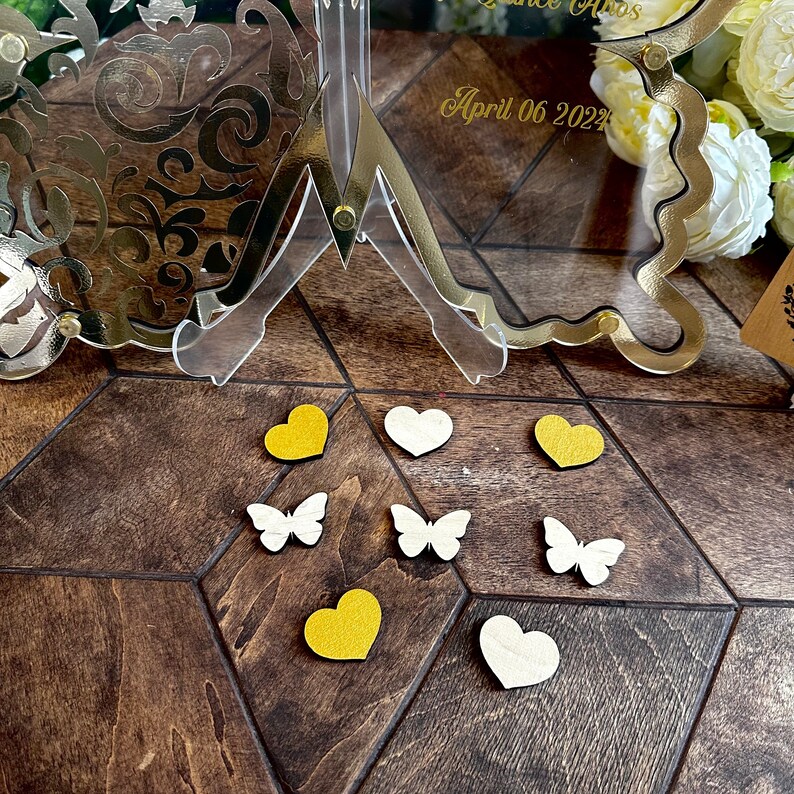 Puede incluir: Una superficie de madera con un soporte decorativo dorado con un marco de vidrio transparente. El marco tiene el texto "April 06 2022" en &eacute;l. Hay varias peque&ntilde;as decoraciones en forma de coraz&oacute;n blancas y amarillas y decoraciones en forma de mariposa en la superficie.