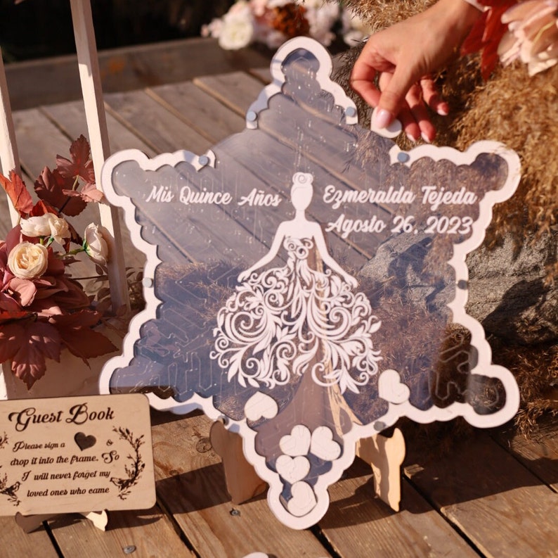 Pu&ograve; includere: Un cartello decorativo a forma di fiocco di neve con un centro trasparente e un bordo bianco. Il cartello presenta un disegno tagliato al laser di un vestito e il testo "Mis Quince A&ntilde;os, Ezmeralda Tejeda, Agosto 26, 2023". &Egrave; visibile anche un piccolo cartello di legno con la scritta "Guest Book".