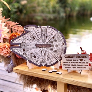 Könnte beinhalten: Ein Gästebuch aus Holz in Form des Millennium Falcon aus Star Wars. Das Gästebuch hat ein Schild mit der Aufschrift "Gästebuch, Bitte ein Herz unterschreiben und in den Rahmen legen, damit wir unsere Lieben, die gekommen sind, nie vergessen."