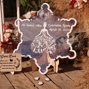 Pu&ograve; includere: Un cartello decorativo a forma di fiocco di neve con il testo "Mis Quince A&ntilde;os" e "Ezmeralda Tejeda Agosto 26, 2023". Il cartello presenta un disegno di abito tagliato al laser ed &egrave; esposto su una superficie di legno.
