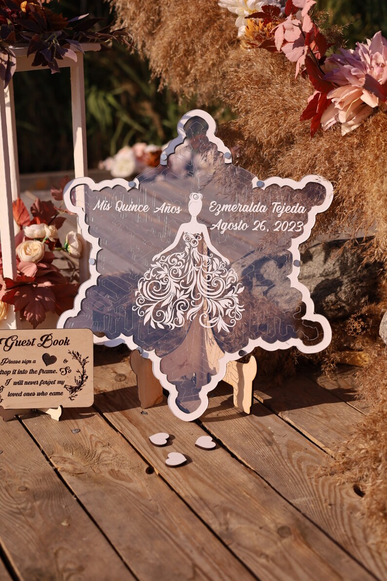 Pu&ograve; includere: Un cartello decorativo a forma di fiocco di neve con il testo "Mis Quince A&ntilde;os" e "Ezmeralda Tejeda Agosto 26, 2023". Il cartello presenta un disegno di abito tagliato al laser ed &egrave; esposto su una superficie di legno.