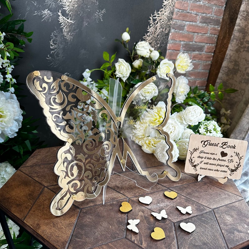 Puede incluir: Un libro de visitas de mariposa de acr&iacute;lico transparente con un dise&ntilde;o de filigrana dorada. La mariposa est&aacute; de pie sobre una mesa de madera con corazones blancos y dorados y mariposas esparcidas a su alrededor. Tambi&eacute;n hay un letrero de madera en la mesa con el texto "Libro de visitas" y "Por favor, firme un trozo y col&oacute;quelo en el marco. As&iacute;, nunca olvidar&eacute; a mis seres queridos que vinieron".
