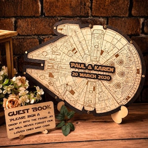 May include: A wooden guest book shaped like a spaceship with engraved details, featuring the text PAUL & KAREN and 20 MARCH 2020. A separate wooden sign reads GUEST BOOK, PLEASE SIGN A HEART & DROP IT INTO THE FRAME. SO WE WILL NEVER FORGET OUR LOVED ONES WHO CAME.