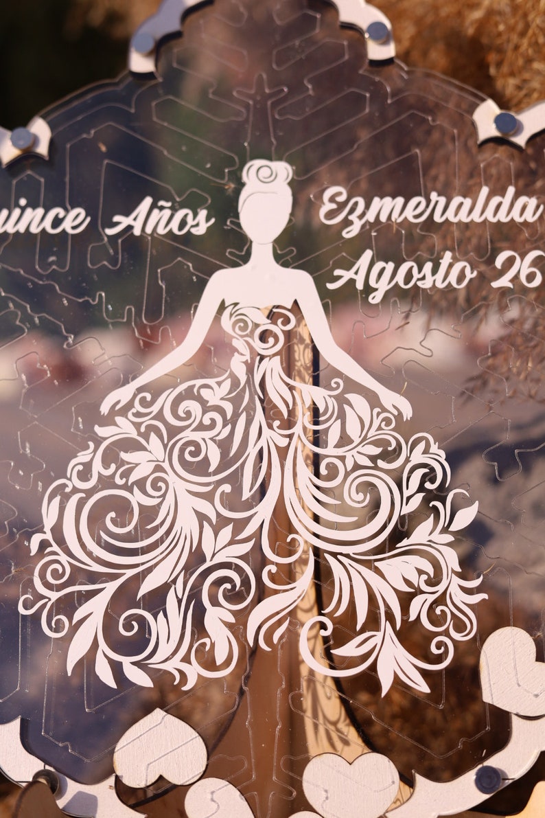Pu&ograve; includere: Un pezzo decorativo in acrilico trasparente a forma di fiocco di neve. Presenta una silhouette ritagliata di una donna in abito con intricati disegni floreali. &Egrave; visibile anche il testo "Quince A&ntilde;os" e "Ezmeralda Agosto 26".