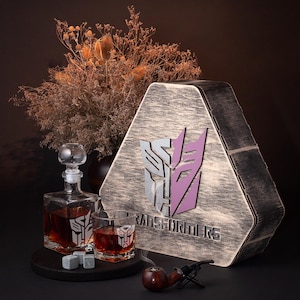 May include: A wooden box with a triangular shape and a Transformers logo. The box is decorated with a distressed wood finish. The box includes a glass decanter, two glasses, and a pipe.