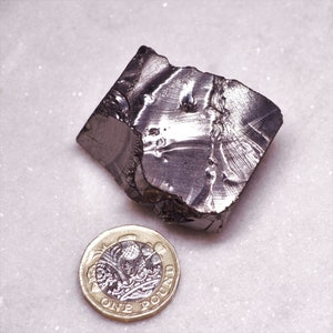 May include: A large, irregularly shaped piece of dark gray metal with a shiny, metallic finish. The metal is next to a British one pound coin for scale.