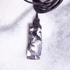 May include: A black cord necklace with a silver-colored, rectangular pendant made of meteorite. The pendant has a rough, textured surface.