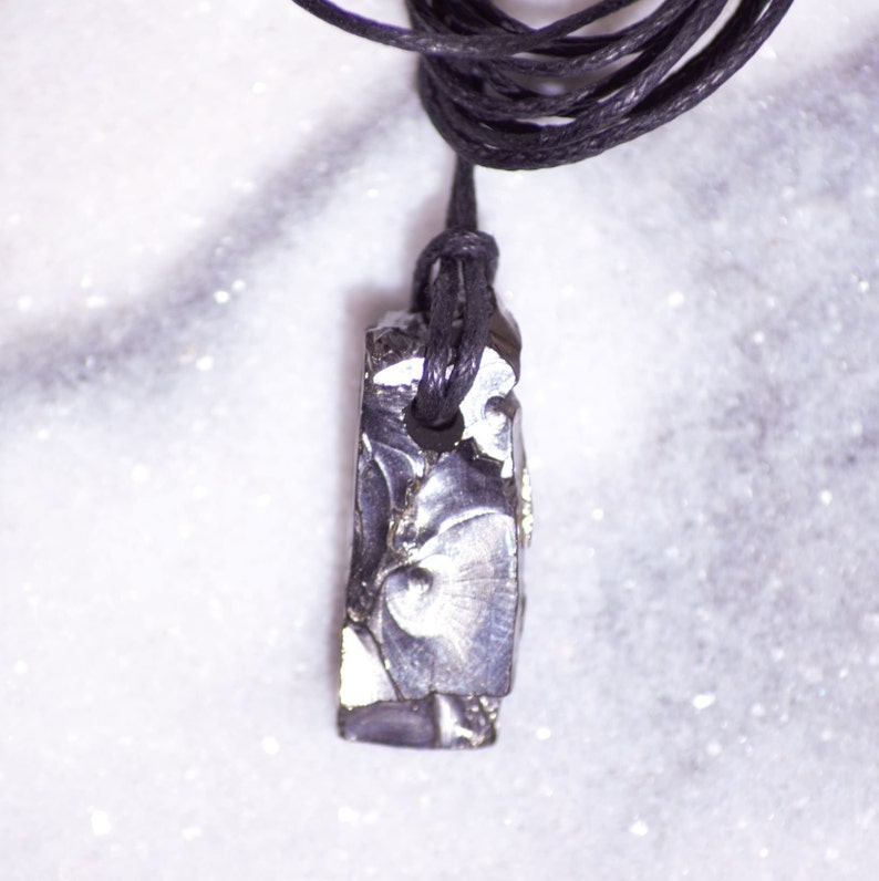 May include: A black cord necklace with a silver-colored, rectangular pendant made of meteorite. The pendant has a rough, textured surface.