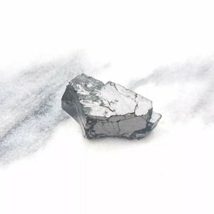 May include: A dark gray, rough, and irregularly shaped piece of raw, unpolished, natural stone. The stone has a metallic sheen and is sitting on a white surface.