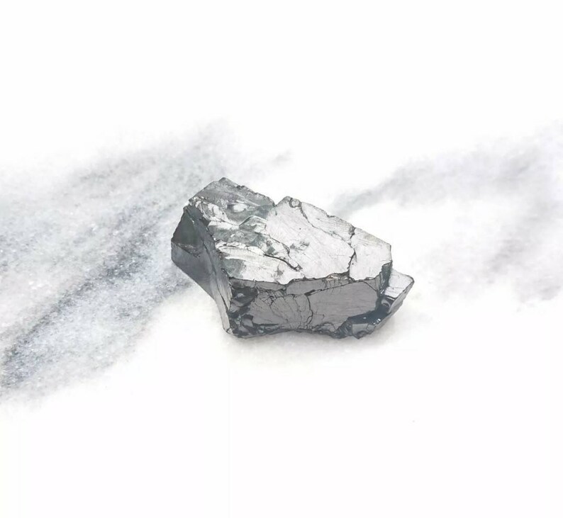 May include: A dark gray, rough, and irregularly shaped piece of raw, unpolished, natural stone. The stone has a metallic sheen and is sitting on a white surface.