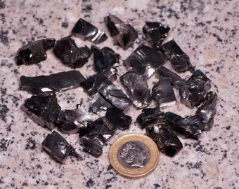 Könnte beinhalten: Eine Ansammlung unregelmäßig geformter, glänzender schwarzer Obsidian-Splitter auf einer gesprenkelten Oberfläche. Eine goldfarbene Münze mit Porträt dient als Größenvergleich. Die Obsidianstücke sind unterschiedlich groß.