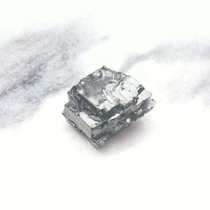 May include: A close-up of a silvery-gray, metallic, irregularly shaped chunk of gallium metal. The metal has a crystalline structure and appears to be slightly reflective.