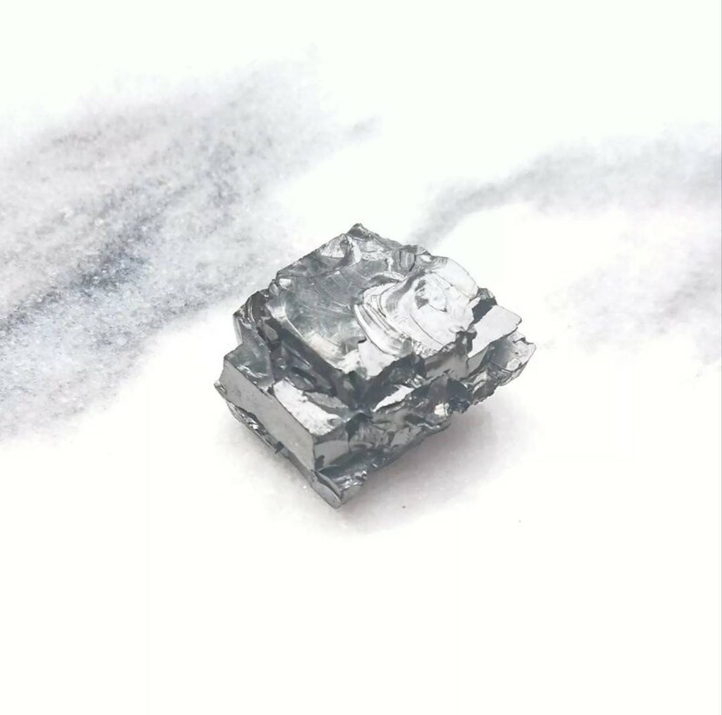 May include: A close-up of a silvery-gray, metallic, irregularly shaped chunk of gallium metal. The metal has a crystalline structure and appears to be slightly reflective.