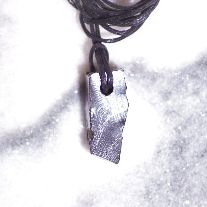 May include: A silver-colored pendant with a rough, irregular shape hangs from a black cord. The pendant is attached to the cord with a small loop.