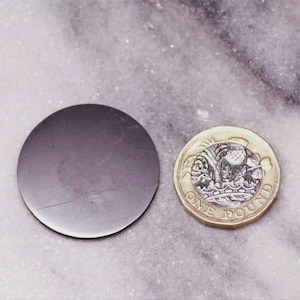 May include: A dark grey, circular object with a cracked surface is next to a British one-pound coin. The coin is gold and silver in colour, with intricate designs and the words "ONE POUND".