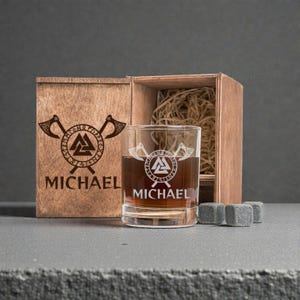 May include: A personalized whiskey glass with a Viking-themed design, filled with amber liquid, sits next to a wooden box with matching engravings. The box contains decorative straw. Four gray whiskey stones are in front of the glass. The name "MICHAEL" is engraved on the glass and box.