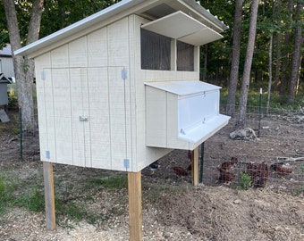 May include: A white wooden chicken coop with a slanted roof and a nesting box. The coop is raised on four wooden posts and is surrounded by a green wire fence. There are several brown chickens in the fenced area.