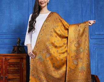 May include: A mustard-yellow shawl with detailed floral and paisley designs. The shawl features a fringed border and is worn by a person in a white outfit. The backdrop is a plain blue wall.