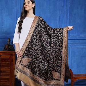 Puede incluir: Un chal negro con un intrincado diseño floral y un borde beige. El chal está drapeado, mostrando el diseño detallado. El fondo es una pared azul sólida.