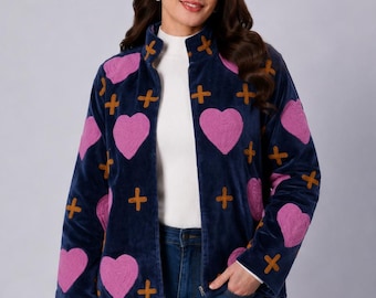 May include: A navy blue zip-up jacket featuring a repeating pattern of embroidered pink hearts and mustard yellow plus signs. The jacket has a high collar, long sleeves, and a straight hemline, worn over a white mock neck top and blue jeans.