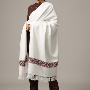 May include: A white shawl with a brown and pink embroidered border and fringe. The shawl is draped over a person wearing a brown top and pants.