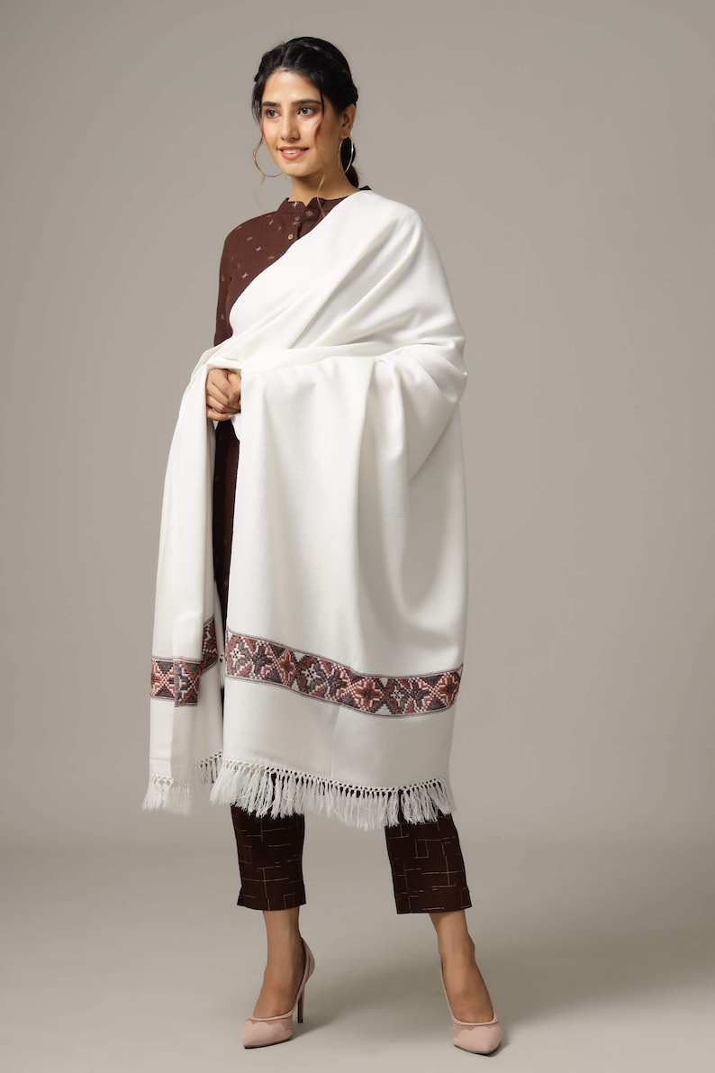 May include: A white shawl with a brown and pink embroidered border and fringe. The shawl is draped over a person wearing a brown top and pants.