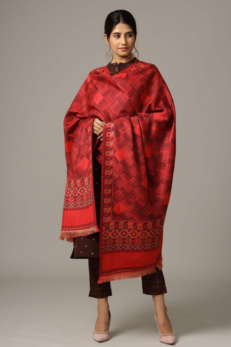 May include: A red shawl with a geometric pattern and a paisley border. The shawl is draped over a person wearing a brown outfit.