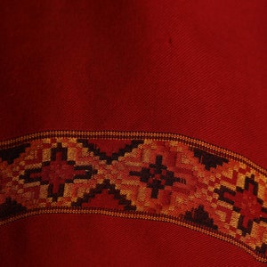 May include: A red fabric with a woven border in black, orange, and gold. The border features a geometric pattern.