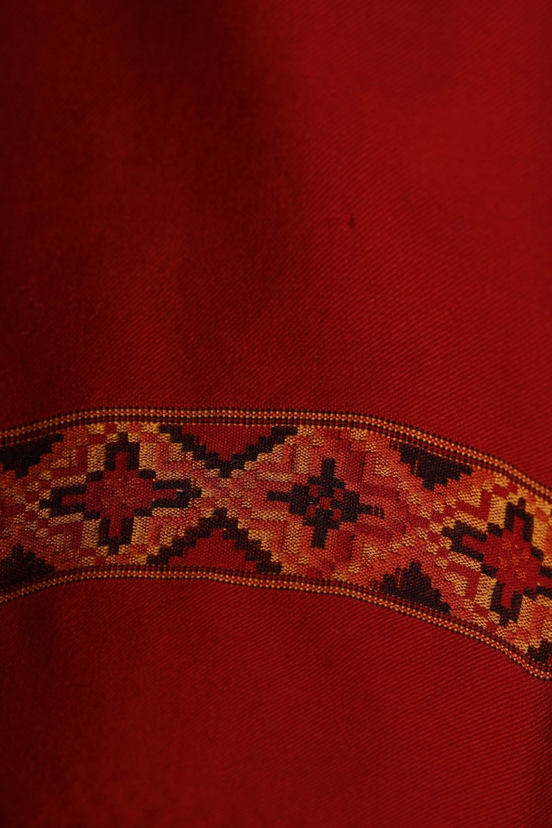 May include: A red fabric with a woven border in black, orange, and gold. The border features a geometric pattern.